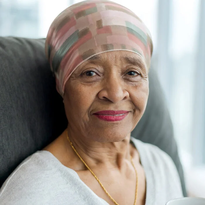 A black senior woman with cancer is wearing a scarf on her head. She is sitting at home in her living room. She is smiling thoughtfully at the camera. She is full of gratitude and hope for recovery.