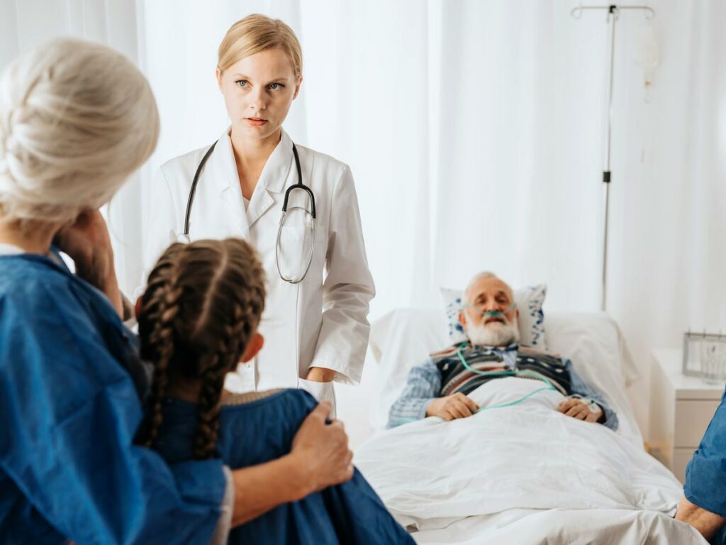 Homebound patient in bed, doctor discussing end-of-life care with spouse and granddaughter during a difficult conversation.