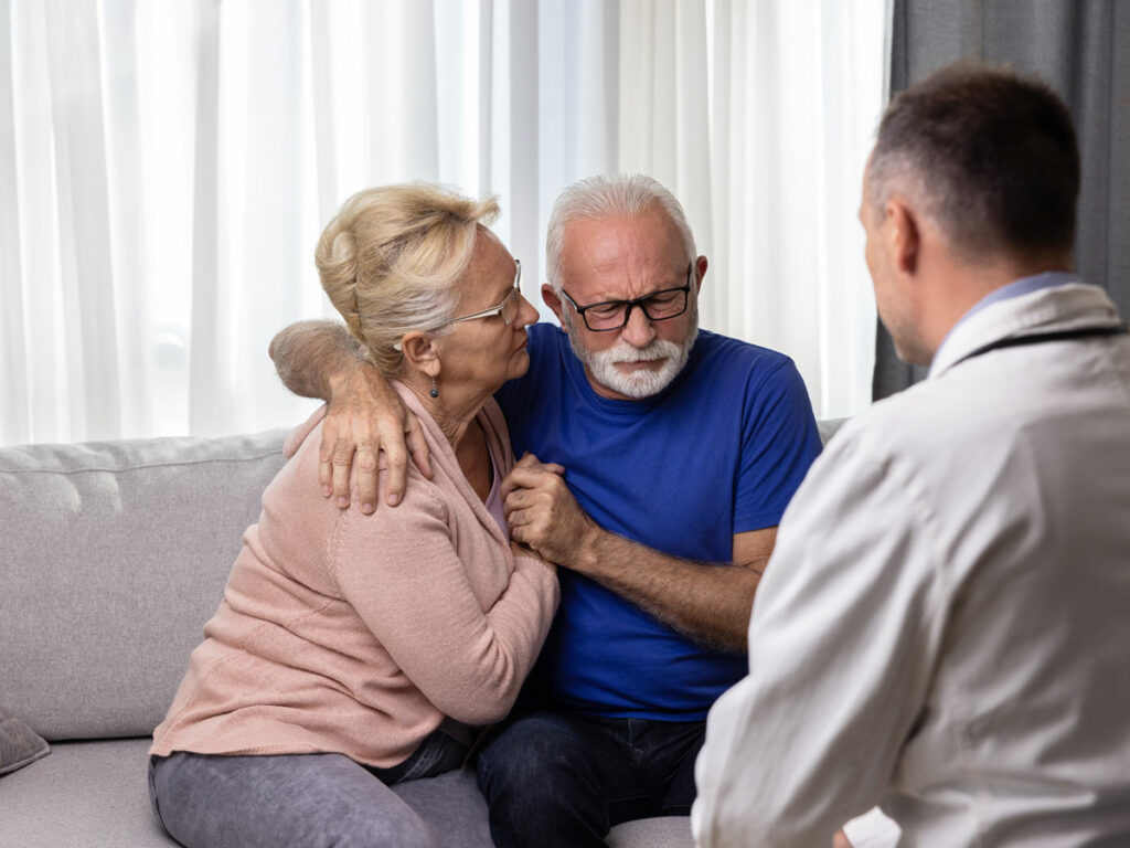 A compassionate doctor delivers a terminal diagnosis to a visibly upset male patient, while his wife provides comforting support through a heartfelt embrace. A moment of empathy and understanding in difficult times.