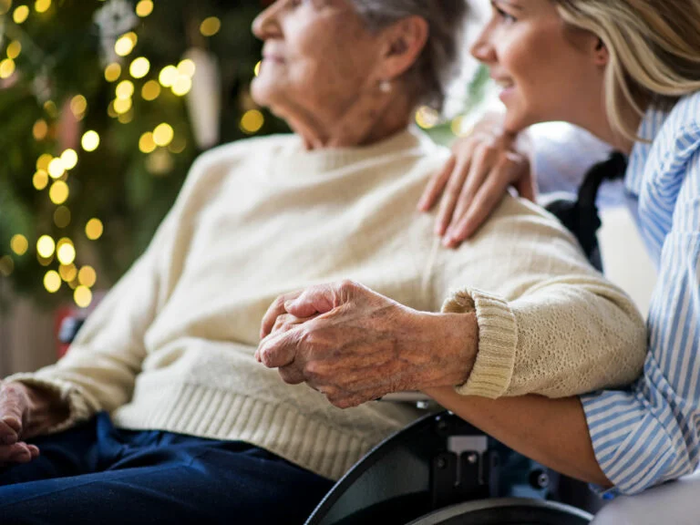 An elderly woman in a wheelchair is tenderly hugged by a caretaker in a Christmas-decorated group home. Festive lights and a tree add warmth to the scene.