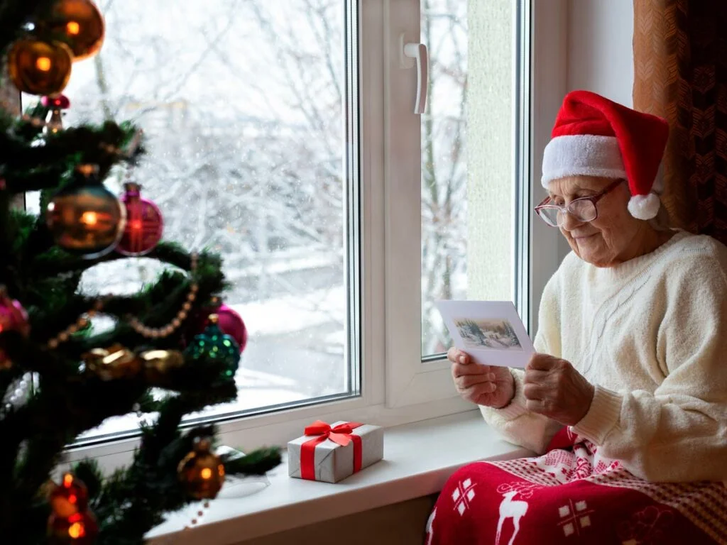 Elderly Caucasian woman in respite care, wearing a Santa hat and glasses, sits by a window. She is intently reading a greeting card, her face illuminated by natural light.