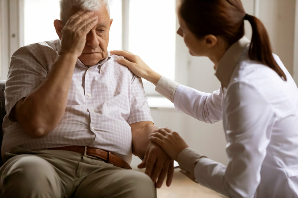 Palliative Care patient holding his head in pain while a nurse holds his hand.