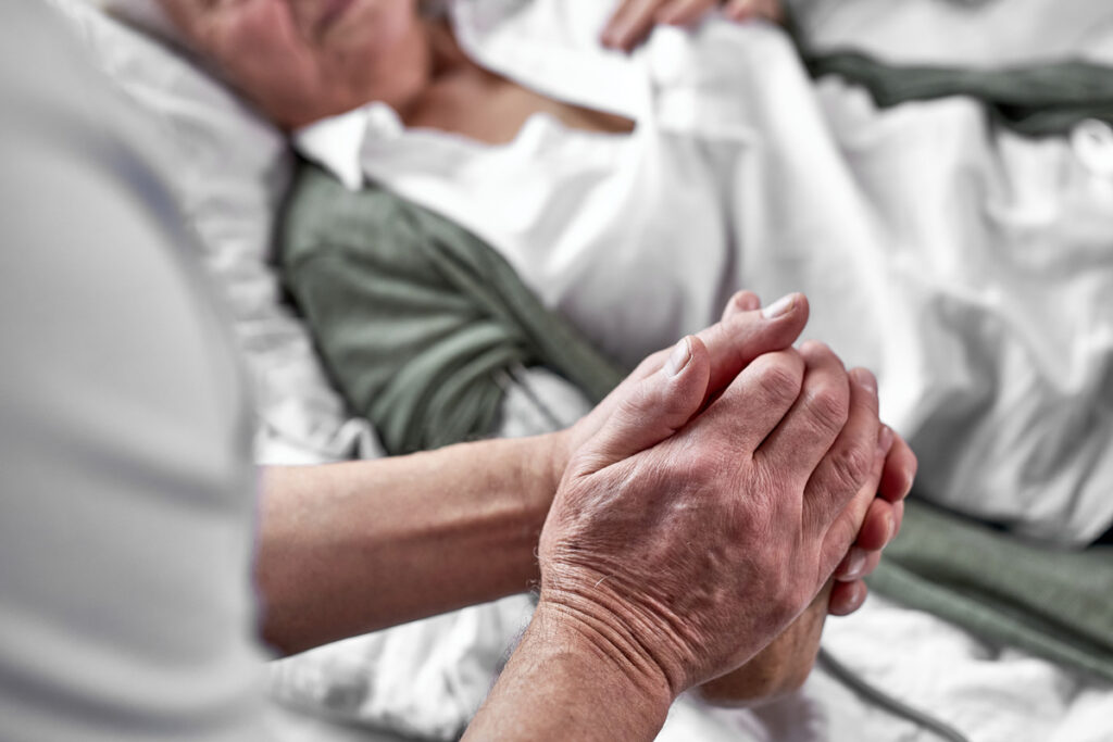 Dying female hospice patient in bed while loved one holds her hand.