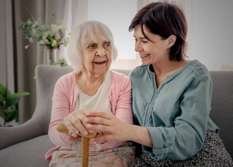 Female hospice volunteer visiting a female patient. Both are sitting on a couch talking and smiling, in the patient's home.